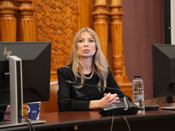 Woman with long blonde hair sits at a desk with a microphone, flanked by two computer monitors in a formal room.