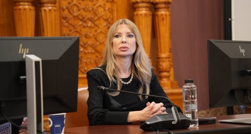 Woman with long blonde hair sits at a desk with a microphone, flanked by two computer monitors in a formal room.