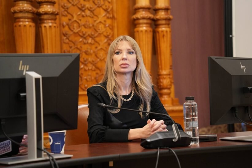 Woman with long blonde hair sits at a desk with a microphone, flanked by two computer monitors in a formal room.