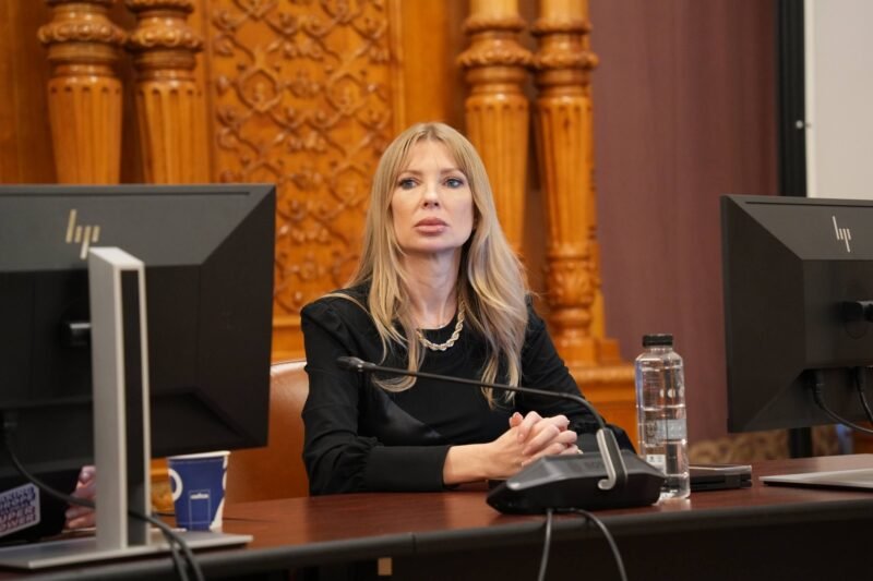Woman with long blonde hair sits at a desk with a microphone, flanked by two computer monitors in a formal room.