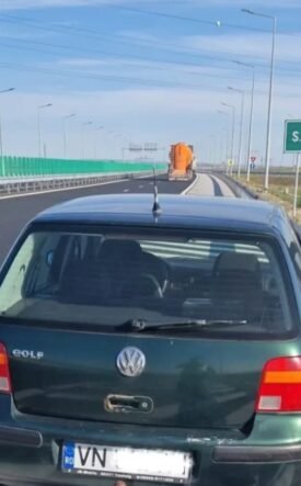 Green VW Golf pulled over on a highway shoulder; an elderly man in a brown jacket stands by the open driver's door beside a metal guardrail, under a clear blue sky with an SOS 112 sign visible in the distance.