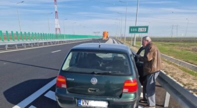 Green VW Golf pulled over on a highway shoulder; an elderly man in a brown jacket stands by the open driver's door beside a metal guardrail, under a clear blue sky with an SOS 112 sign visible in the distance.