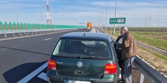 Green VW Golf pulled over on a highway shoulder; an elderly man in a brown jacket stands by the open driver's door beside a metal guardrail, under a clear blue sky with an SOS 112 sign visible in the distance.