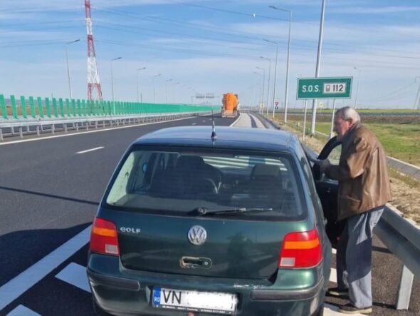 Green VW Golf pulled over on a highway shoulder; an elderly man in a brown jacket stands by the open driver's door beside a metal guardrail, under a clear blue sky with an SOS 112 sign visible in the distance.