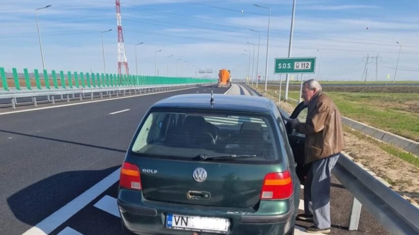 Green VW Golf pulled over on a highway shoulder; an elderly man in a brown jacket stands by the open driver's door beside a metal guardrail, under a clear blue sky with an SOS 112 sign visible in the distance.
