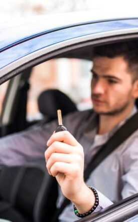 Man in a car holding up a car key toward the camera while wearing a seatbelt in a blue vehicle.