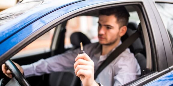 Man in a car holding up a car key toward the camera while wearing a seatbelt in a blue vehicle.