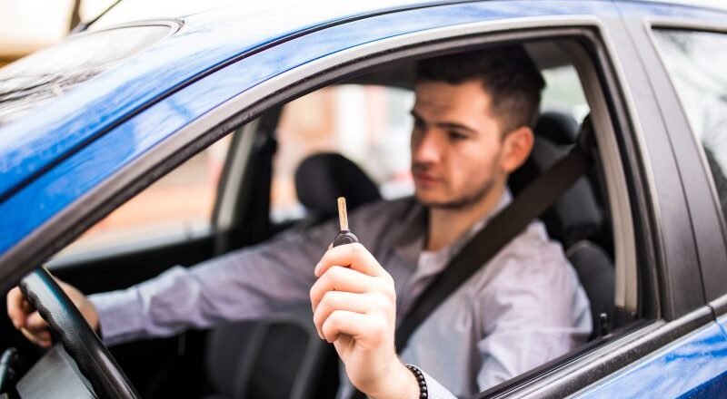 Man in a car holding up a car key toward the camera while wearing a seatbelt in a blue vehicle.