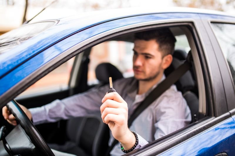 Man in a car holding up a car key toward the camera while wearing a seatbelt in a blue vehicle.