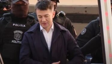 A man in a white shirt and dark jacket stands surrounded by masked police officers labeled POLITIA outside a vehicle area.