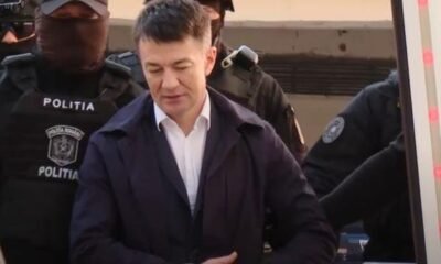 A man in a white shirt and dark jacket stands surrounded by masked police officers labeled POLITIA outside a vehicle area.