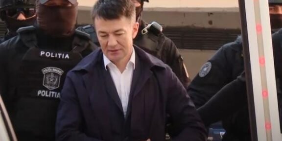 A man in a white shirt and dark jacket stands surrounded by masked police officers labeled POLITIA outside a vehicle area.