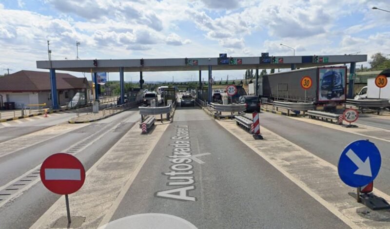 Toll plaza on a highway with multiple lanes, barriers, and cars waiting to pass under an overhead canopy.