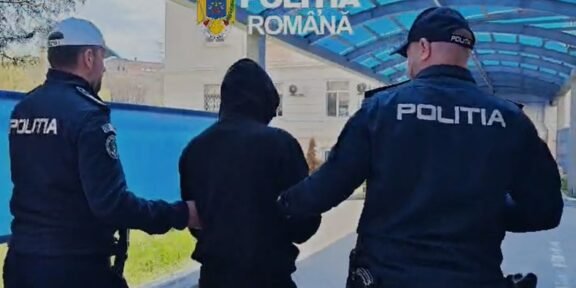 Two Romanian police officers escort a hooded person outdoors under a blue canopy labeled Politia Romana.