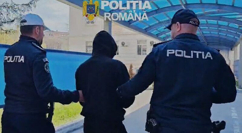 Two Romanian police officers escort a hooded person outdoors under a blue canopy labeled Politia Romana.