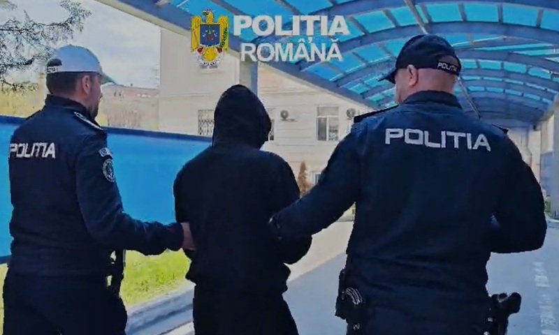 Two Romanian police officers escort a hooded person outdoors under a blue canopy labeled Politia Romana.