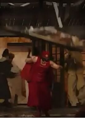 People rush through a doorway as debris falls from a damaged interior during an explosion scene, with a person in red clothing center-stage guarding the exit.