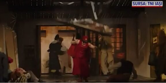 People rush through a doorway as debris falls from a damaged interior during an explosion scene, with a person in red clothing center-stage guarding the exit.