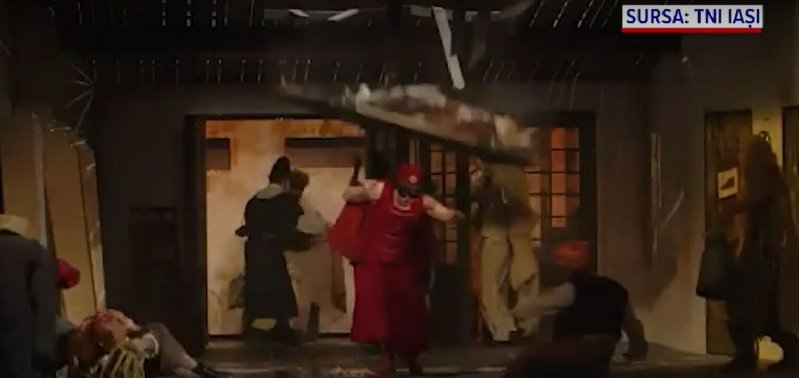 People rush through a doorway as debris falls from a damaged interior during an explosion scene, with a person in red clothing center-stage guarding the exit.