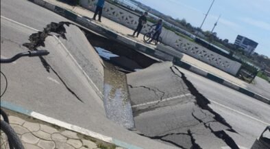 A wide street sinkhole with cracked asphalt, filled with water, spilling toward the curb while three people and a cyclist observe from the sidewalk behind a decorative railing.