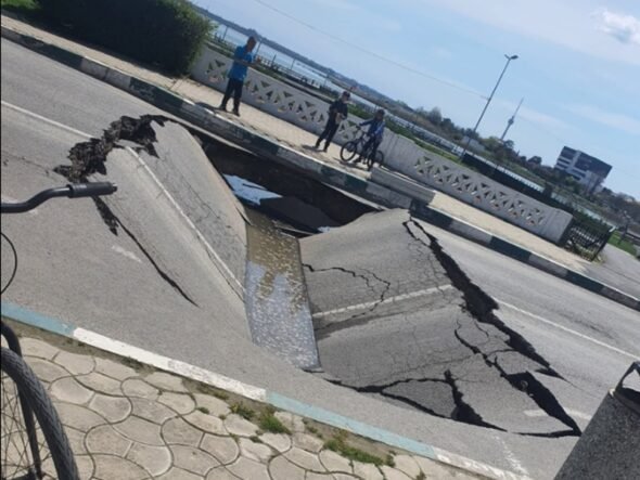 A wide street sinkhole with cracked asphalt, filled with water, spilling toward the curb while three people and a cyclist observe from the sidewalk behind a decorative railing.