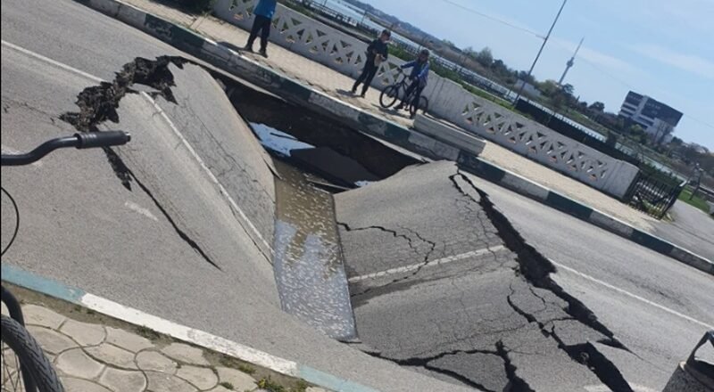 A wide street sinkhole with cracked asphalt, filled with water, spilling toward the curb while three people and a cyclist observe from the sidewalk behind a decorative railing.