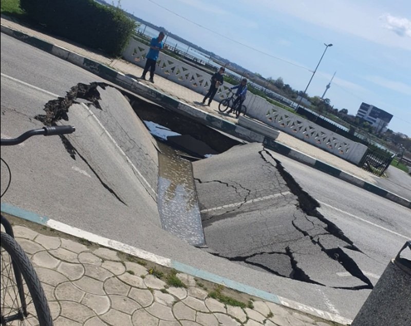 A wide street sinkhole with cracked asphalt, filled with water, spilling toward the curb while three people and a cyclist observe from the sidewalk behind a decorative railing.