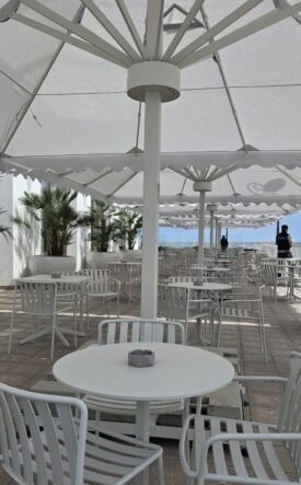 Outdoor seaside cafe with white tables and chairs under large white shade umbrellas along a railing overlooking the ocean.