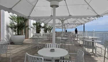 Outdoor seaside cafe with white tables and chairs under large white shade umbrellas along a railing overlooking the ocean.