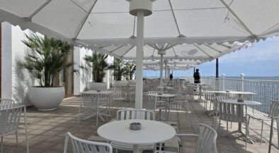 Outdoor seaside cafe with white tables and chairs under large white shade umbrellas along a railing overlooking the ocean.