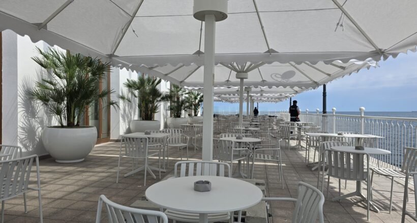 Outdoor seaside cafe with white tables and chairs under large white shade umbrellas along a railing overlooking the ocean.