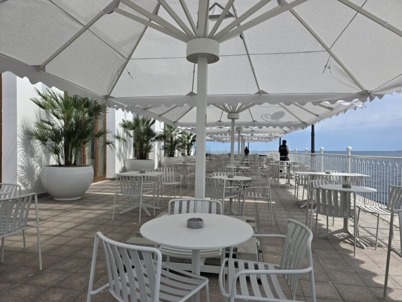 Outdoor seaside cafe with white tables and chairs under large white shade umbrellas along a railing overlooking the ocean.