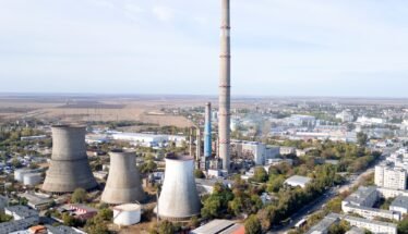 Aerial view of a large industrial plant with tall smokestack and several cooling towers near a town horizon