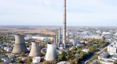 Aerial view of a large industrial plant with tall smokestack and several cooling towers near a town horizon