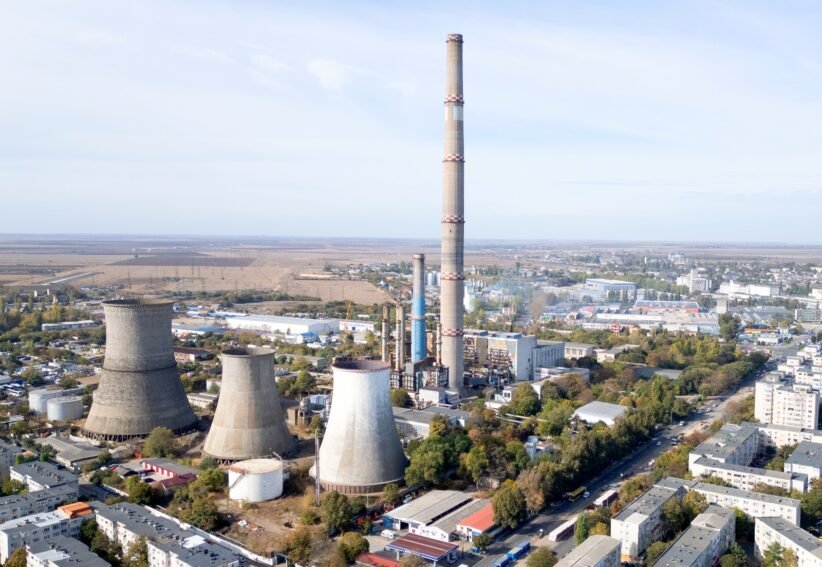 Aerial view of a large industrial plant with tall smokestack and several cooling towers near a town horizon