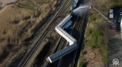 Aerial view of several derailed train cars on tracks; firefighters spray water from a ladder truck at the scene.