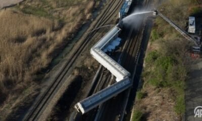 Aerial view of several derailed train cars on tracks; firefighters spray water from a ladder truck at the scene.