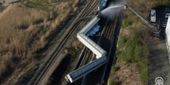 Aerial view of several derailed train cars on tracks; firefighters spray water from a ladder truck at the scene.