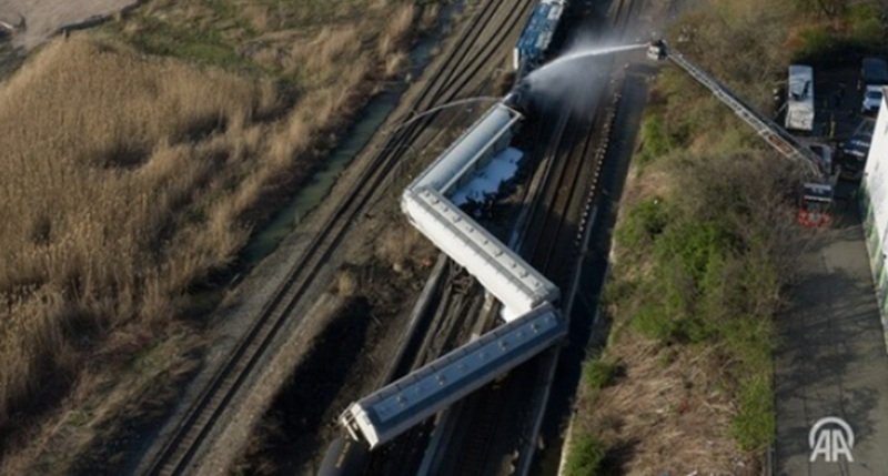 Aerial view of several derailed train cars on tracks; firefighters spray water from a ladder truck at the scene.