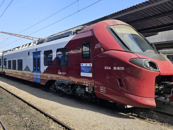Red and white passenger train parked at a platform with overhead electric lines visible.
