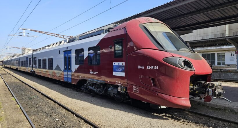 Red and white passenger train parked at a platform with overhead electric lines visible.