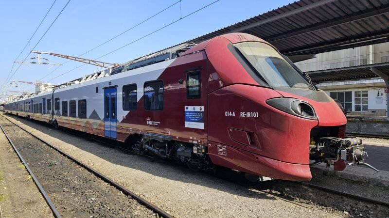 Red and white passenger train parked at a platform with overhead electric lines visible.