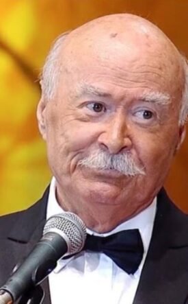 Older man in a black tuxedo and bow tie speaks into a microphone on stage against a warm orange background.