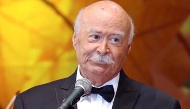 Older man in a black tuxedo and bow tie speaks into a microphone on stage against a warm orange background.