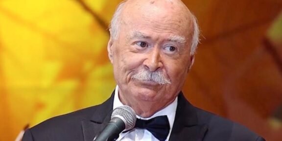 Older man in a black tuxedo and bow tie speaks into a microphone on stage against a warm orange background.