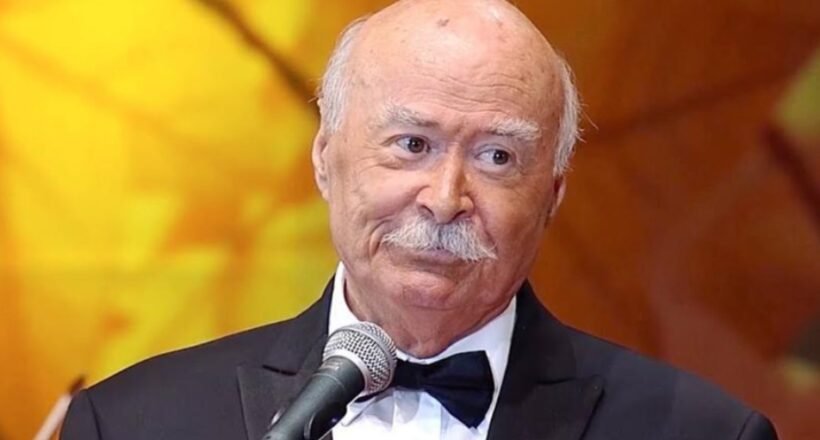 Older man in a black tuxedo and bow tie speaks into a microphone on stage against a warm orange background.