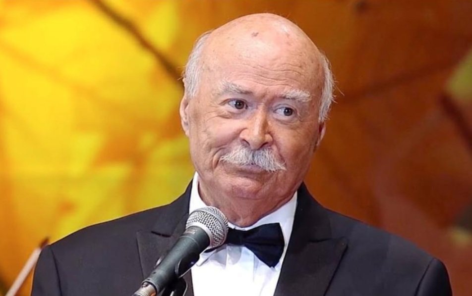 Older man in a black tuxedo and bow tie speaks into a microphone on stage against a warm orange background.