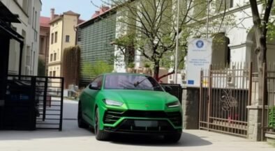 Bright green Lamborghini SUV driving down a gated residential street with parked cars and trees on a sunny day.