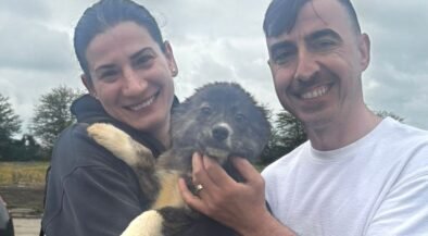 Smiling couple holding a fluffy puppy outdoors against a cloudy skyp