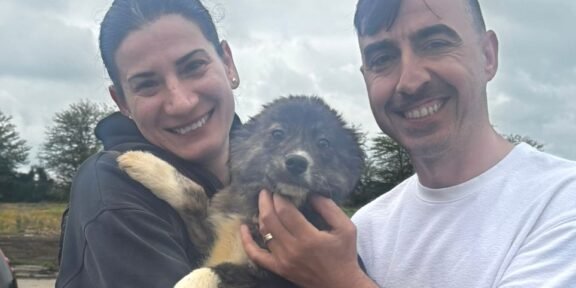 Smiling couple holding a fluffy puppy outdoors against a cloudy skyp